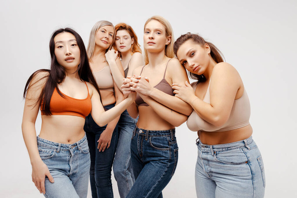 Modern diverse, multiracial group of women wearing sexy bras and jeans hugging, looking at camera. Fashion beautiful models posing in studio, standing isolated on white background. Support concept - Photo, Image