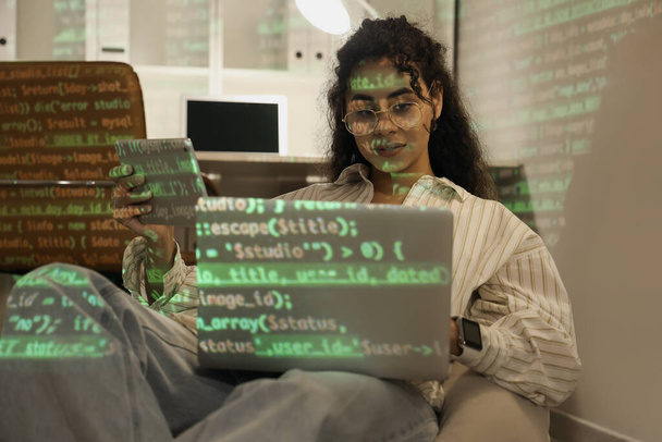 Женщина-афро-американский программист с гаджетами на мешке и проекцией кода в офисе ночью - Фото, изображение