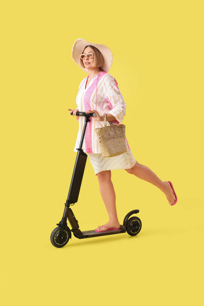 Hermosa mujer feliz madura montando patinete scooter sobre fondo amarillo. Concepto vacaciones de verano - Foto, Imagen
