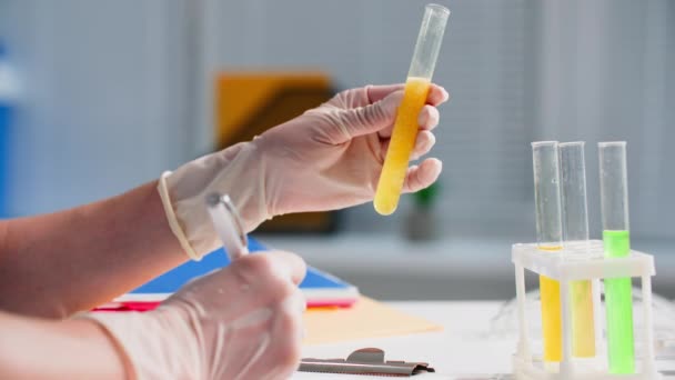 laboratório médico, assistente de laboratório feminino em luvas examina a reação da solução em tubos de ensaio e escreve resultados em um bloco de notas, close-up - Filmagem, Vídeo