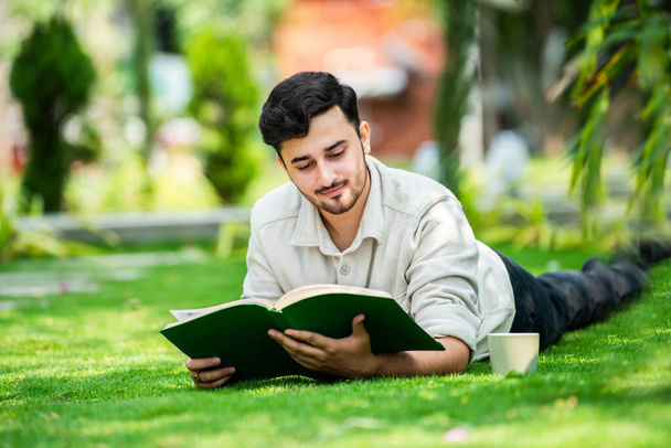 Young attentive Indian male student lies on green grass and reads book at summer green park - Photo, Image