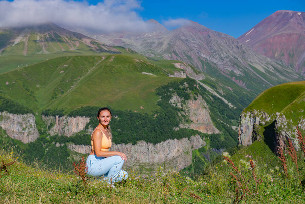 Een mooie vrouw in een stijlvolle blauwe outfit staat in bewondering, kijkend naar het adembenemende uitzicht op een berglandschap met weelderige groene bomen en een helderblauwe lucht op de achtergrond. - Foto, afbeelding