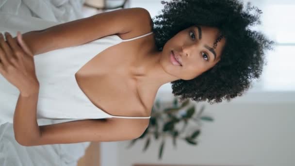 Portrait serene lady sitting bed. Curly brunette woman touching soft skin looking camera alone. Calm dreamy model enjoying lazy morning vertical view. African girl posing at comfortable white bedroom - Footage, Video