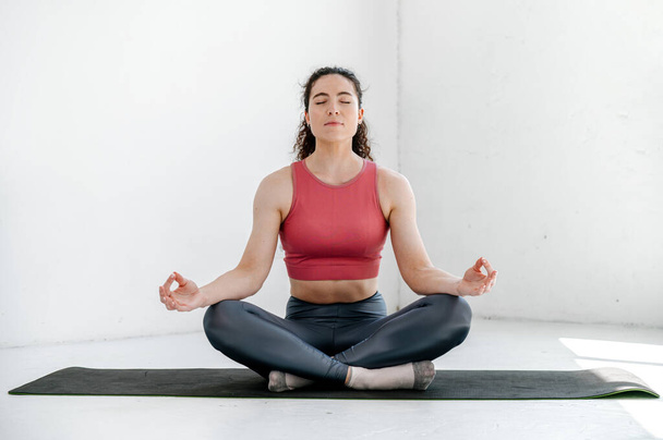 Young woman in sportswear sitting on mat in half lotus pose with mudra gesture, practicing yoga lesson, breathing, meditation, doing exercise, workout. Wellness concept - Photo, Image