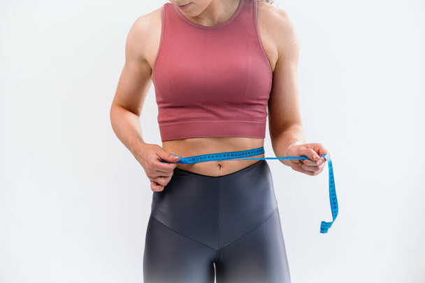 体重とアクティブなライフスタイルを減らすコンセプト. 白い背景に隔離されたウエストラインに沿って青い測定テープを保持している女性のクロップビュー. アクティブウェアチェック結果のスポーティな女性 - 写真・画像