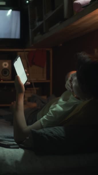 Full vertical shot of sleepy Asian person relaxing on bunk bed in micro apartment, browsing on smartphone, yawning, hugging pillow and lying down, movie playing on TV in background - Footage, Video