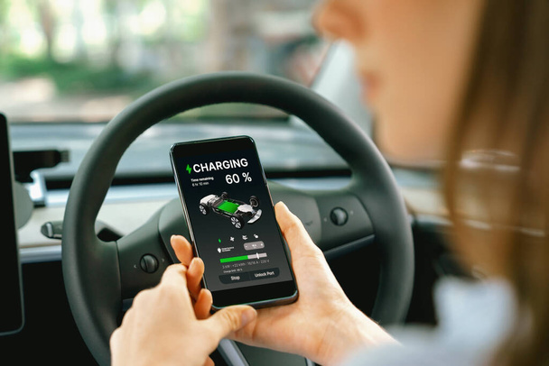 Aracın içindeki genç kadın, modern EV arabasındaki akıllı telefon ekranında akü şarj etme uygulamasını kontrol ediyor. Yükselt - Fotoğraf, Görsel