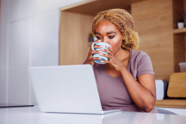 Woman sitting at kitchen counter drinking coffee and using laptop computer while working remotely from home - Foto, Bild