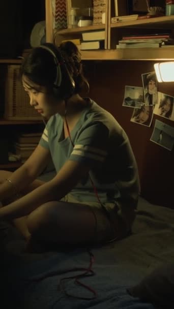 Tiro vertical completo de niña asiática joven sentada en la cama en el dormitorio o dormitorio, jugando juego de disparos en el ordenador portátil en línea, sufriendo la derrota, tirar los auriculares y reaccionar con ira - Imágenes, Vídeo