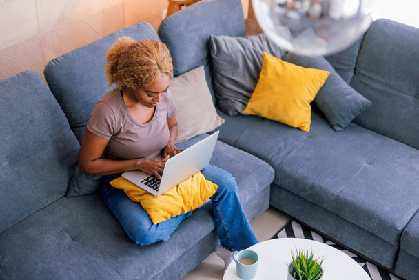High angle view of female freelancer sitting on living room couch working from home using laptop computer and drinking coffee; woman working remotely from home - Photo, Image
