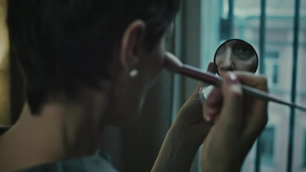 Over shoulder lowkey shot of adult Caucasian woman using makeup to cover bruise under her eye, looking at herself in compact mirror while applying powder on her face in dark cold bedroom - Footage, Video