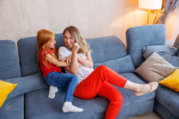 Beautiful mother and little daughter having fun spending leisure time together at home, tickling, cuddling and laughing - Foto, Bild