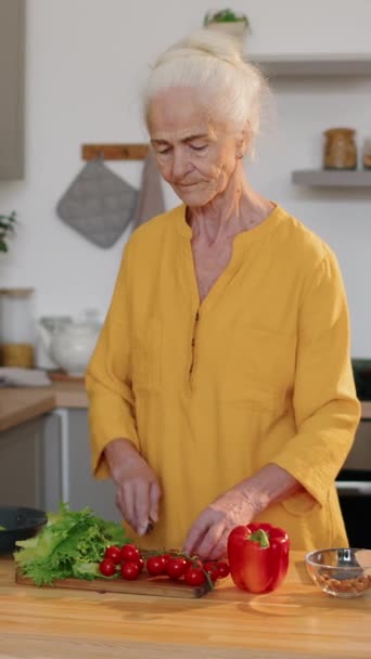 Κάθετη μέτρια βολή της υγιή Καυκάσια ηλικιωμένη γυναίκα φορώντας μακρύ φόρεμα μουστάρδα στέκεται στο τραπέζι μαγειρέματος στην κουζίνα και κοπή φρέσκα λαχανικά, ενώ κάνοντας σαλάτα για μεσημεριανό γεύμα στο σπίτι - Πλάνα, βίντεο