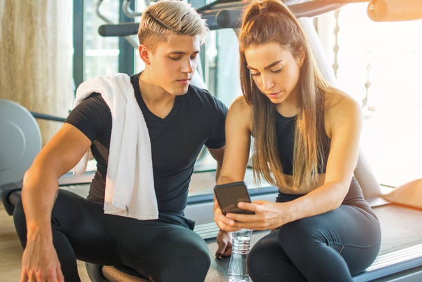 Sportowa para sprawdzająca posty w mediach społecznościowych za pomocą smartfona podczas przerwy na siłownię. Młodzi przyjaciele siedzący na bieżni i korzystający ze wspólnego telefonu na siłowni. - Zdjęcie, obraz