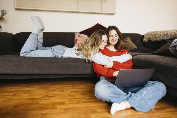 Lesbo pari makaa sohvalla kotona, puhuu hiljaa ja hyväilee toisiaan, kun käytät kannettavaa tietokonetta. Nuori naispari jakaa hellyyden ja kiintymyksen hetken intiimissä ja kodikkaassa ilmapiirissä.. - Valokuva, kuva