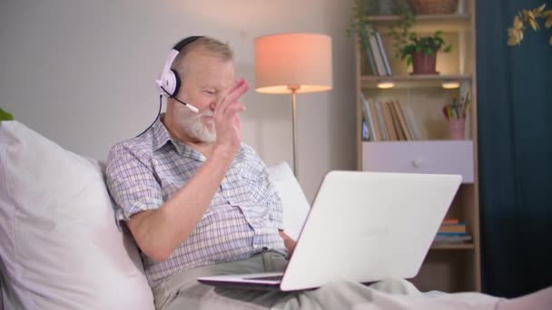 modern nyugdíjas mikrofonnal és fejhallgatóval beszél videokommunikációval egy laptopon, miközben a kanapén ül a szobában - Felvétel, videó