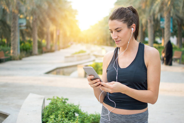 Молодая фитнес-женщина с наушником с помощью смартфона в парке - Фото, изображение