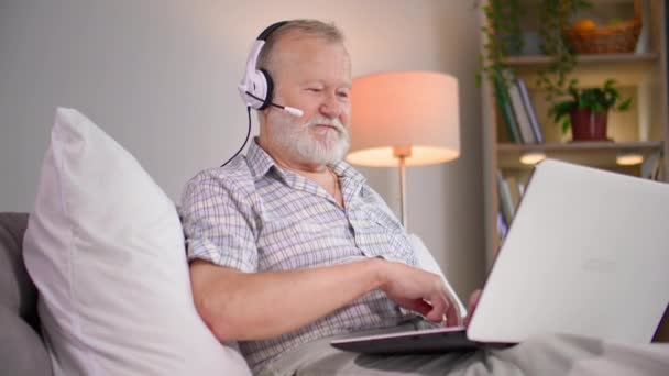 atractivo anciano hablando en videollamada en el portátil usando micrófono y auriculares mientras está sentado en el sofá en el dormitorio - Imágenes, Vídeo