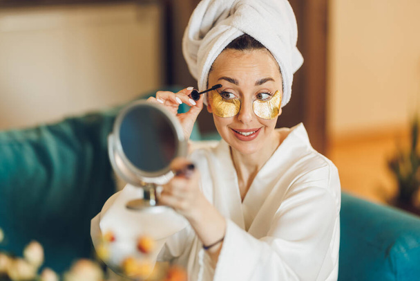 Una donna attraente con la cura della pelle sotto le macchie sugli occhi che applica il mascara mentre si gode la mattina a casa sua. - Foto, immagini