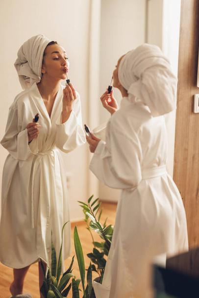 Приваблива жінка в халаті наносить блиск для губ перед великим дзеркалом, насолоджуючись ранком у себе вдома. - Фото, зображення