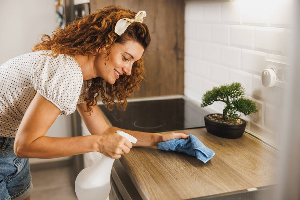 Νεαρή χαμογελαστή γυναίκα κάνει τις καθημερινές δουλειές της στο σπίτι. Καθαρίζει και απολυμαίνει έναν πάγκο κουζίνας.. - Φωτογραφία, εικόνα