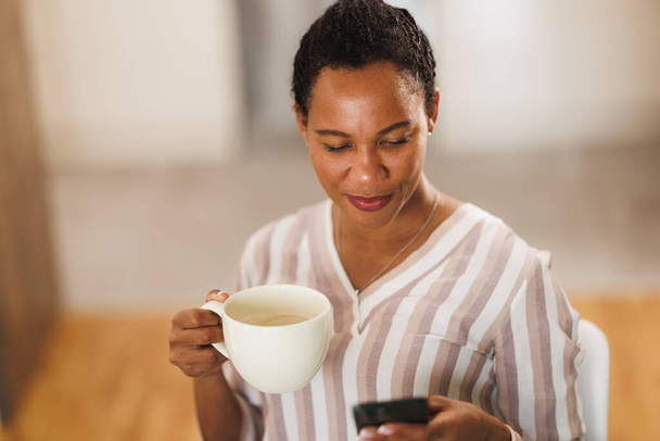 Afrikanische Geschäftsfrau genießt eine Tasse Kaffee und benutzt ein Handy, während sie von zu Hause aus arbeitet. - Foto, Bild