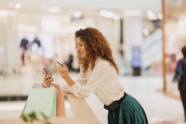 Aufnahme einer lächelnden Frau, die mit ihrem Smartphone Verkaufsangebote online abruft oder Rabatt-App nutzt, während sie in einem Einkaufszentrum einkauft. - Foto, Bild