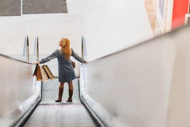 Πίσω όψη μιας χαμογελαστής γυναίκας που στέκεται στις κυλιόμενες σκάλες σε ένα εμπορικό κέντρο. - Φωτογραφία, εικόνα