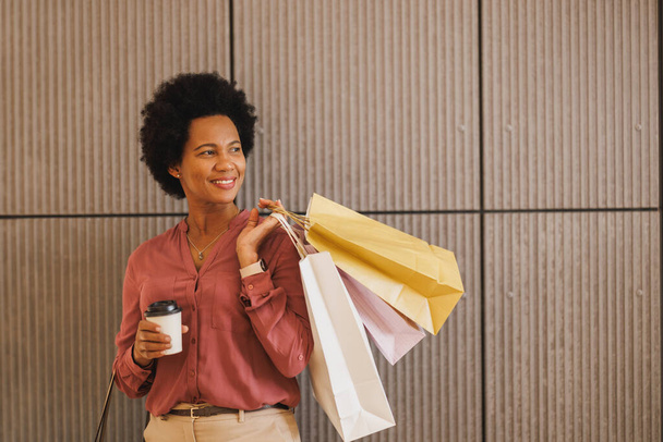 Alışveriş poşetleri taşıyan ve elinde bir fincan kahveyle bir alışveriş merkezinin duvarına dayanmış siyah bir kadının fotoğrafı.. - Fotoğraf, Görsel