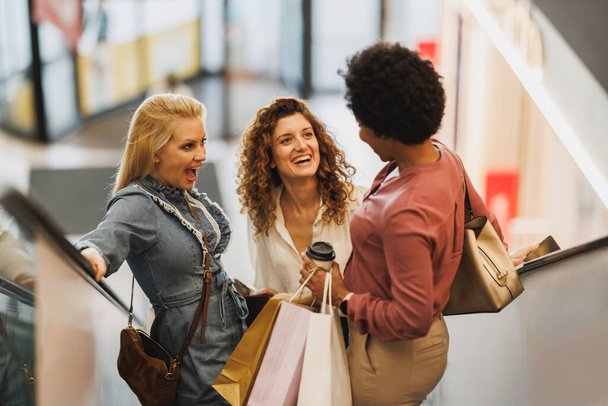 Opname van drie lachende vrouwen die plezier hebben en samen op de roltrap staan in een winkelcentrum terwijl ze aan het winkelen zijn. - Foto, afbeelding