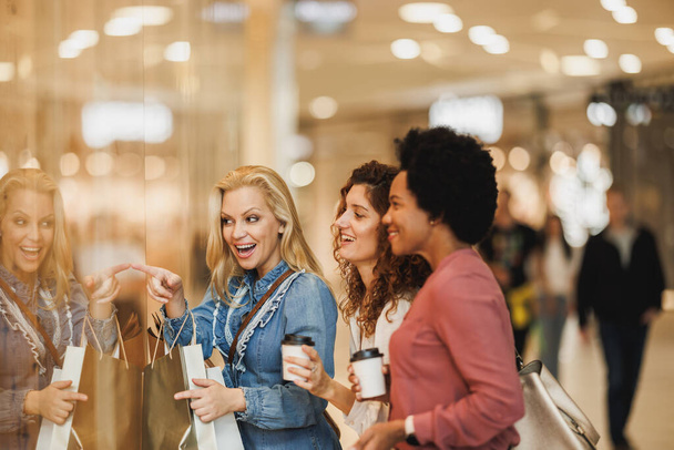 Opname van drie lachende vriendinnen die naar iets kijken in de etalage van een boetiek terwijl ze genieten van een winkeluitbarsting in het winkelcentrum. - Foto, afbeelding