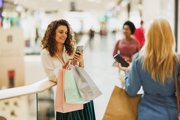 Stijlvolle vrouw met behulp van een mobiele telefoon tijdens het dragen van tassen tijdens een shopping spree in het winkelcentrum. - Foto, afbeelding