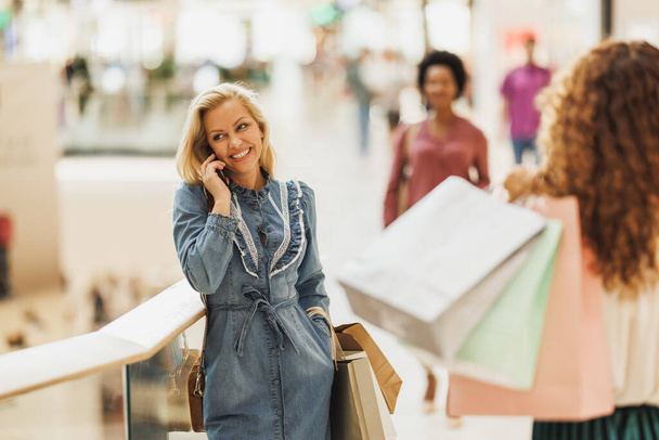Portret van een zeer gelukkige vrouw met behulp van haar smartphone en glimlachen terwijl het houden van boodschappentassen in het winkelcentrum. - Foto, afbeelding