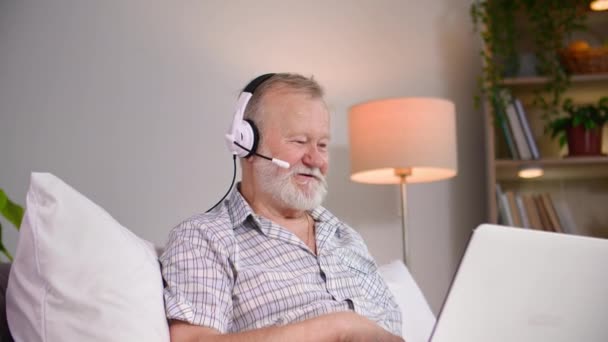 charmant homme âgé dans les écouteurs parler à l'aide d'un microphone par vidéoconférence sur ordinateur portable tout en étant assis sur le canapé dans une salle confortable - Séquence, vidéo