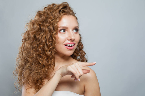 Šokovaná žena ukazuje prstem. Krásná fena model s přirozeným make-up, kudrnaté dlouhé vlasy a přátelský úsměv na bílém pozadí - Fotografie, Obrázek