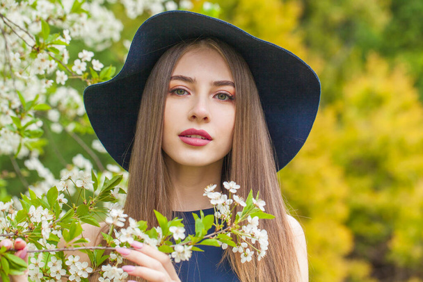 Elegante retrato de mujer morena. Hermosa modelo femenina con pelo largo y maquillaje con sombrero azul clásico en el jardín de primavera al aire libre - Foto, imagen