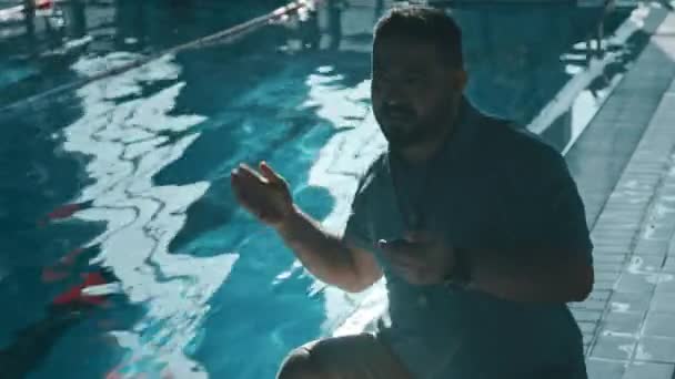 Medium shot with lens flare of Asian male coach crouching by side of pool, cheering at athlete in water, doing crawl stroke arm motions, looking at stopwatch, then praising and showing thumb up - Footage, Video