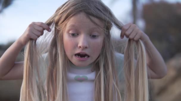 Lustiges kleines Mädchen zieht ihre Haare vor Schreck über das, was sie sieht. Das kleine Mädchen mit den langen blonden Haaren hält spielerisch ihre Haare in den Händen und zeigt eine glückliche Geste. Sie hat Spaß draußen auf der - Filmmaterial, Video