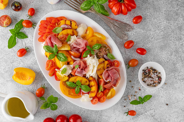 Σαλάτα Caprese με μοτσαρέλα, ντομάτες, προσούτο και ψητά ροδάκινα σε λευκό πιάτο σε γκρι μπετόν φόντο. Φρέσκια καλοκαιρινή σαλάτα. Άνω όψη - Φωτογραφία, εικόνα