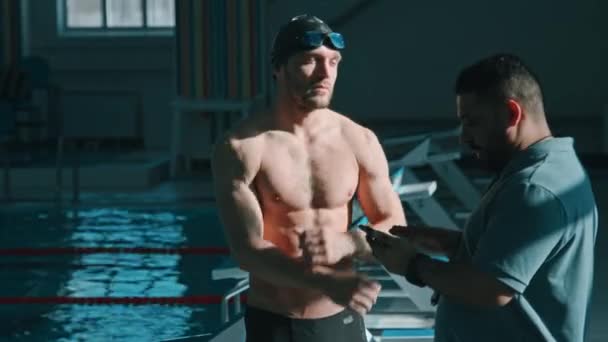 Medium shot van middelbare leeftijd Aziatische man zwemmen coach uit te leggen trainingsplan, het instellen van de praktijk doelen voor jonge atleet, die aandachtig luistert, knikken, opwarmen van de armen en het opzetten van een bril - Video