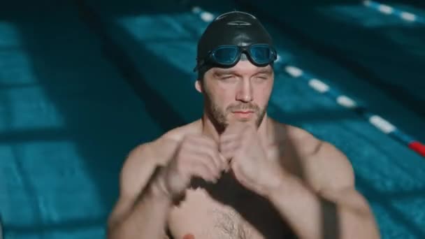 Retrato de close-up médio de jovem nadador masculino caucasiano musculoso fazendo exercícios de aquecimento na piscina antes do treino, colocando óculos e olhando para a câmera - Filmagem, Vídeo