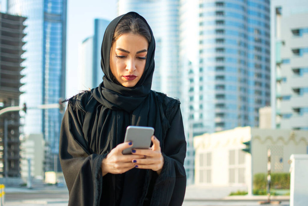 Güzel bir Arap iş kadınının portresi tesettür takıyor. Arka planda gökdelenlerle sokakta cep telefonu kullanıyor. - Fotoğraf, Görsel
