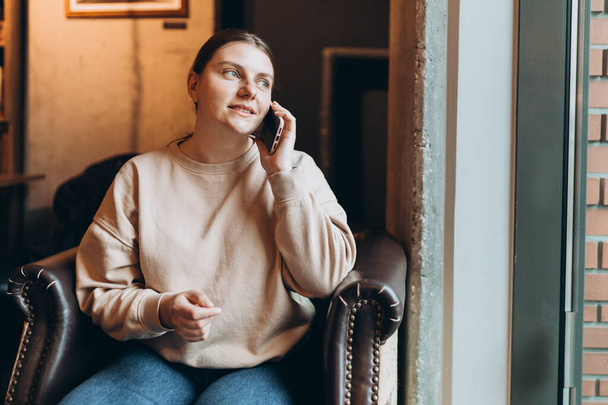 30 'lu yaşlardaki çekici bir kadın kafede dinlenirken cep telefonuyla konuşuyor. Kapalı alanda cep telefonu kullanan sıradan bir genç kadın. - Fotoğraf, Görsel