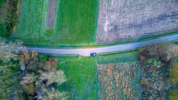 Deze luchtfoto legt een solitair voertuig vast dat over een kronkelende landweg rijdt, omgeven door de lappendeken van velden en bomen die kenmerkend zijn voor het platteland. De rust van de scène - Foto, afbeelding