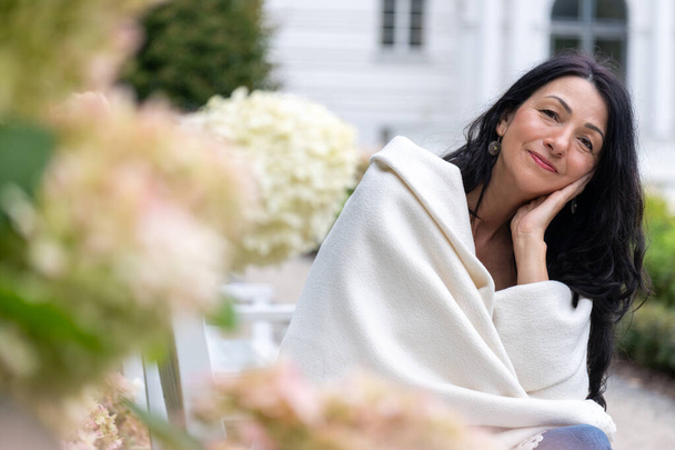 Une femme enveloppée dans un châle crème est assise au milieu des fleurs, son sourire de contenu reflétant l'acceptation de soi et la grâce au milieu de la vie au milieu de la transition hormonale. Photo de haute qualité - Photo, image