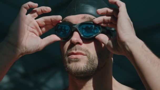Close-up portrait shot of calm, confident young Caucasian male athlete with stubble and beard, in black rubber cap putting on goggles and looking away, while getting ready for competition in pool - Footage, Video