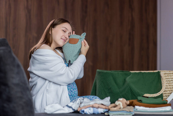 Pregnant woman folding baby clothes sitting on sofa at home. - Zdjęcie, obraz