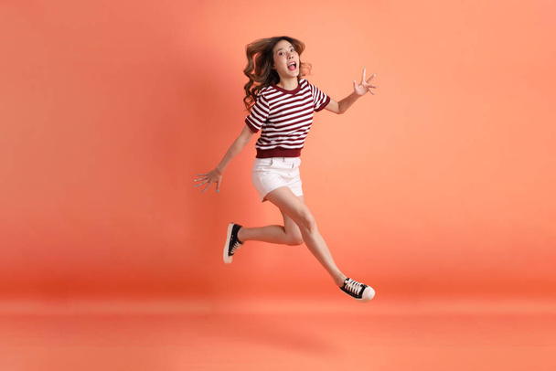 La giovane donna asiatica in abiti casual con gesto di salto sullo sfondo arancione  - Foto, immagini