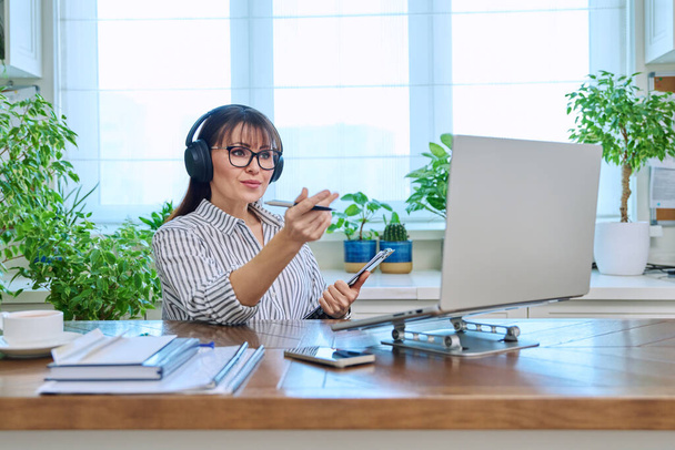 Mature woman in headphones having video conference using laptop computer in home office. Remote virtual meeting, online training, consulting, testing. Work, education, technology, people concept - Φωτογραφία, εικόνα