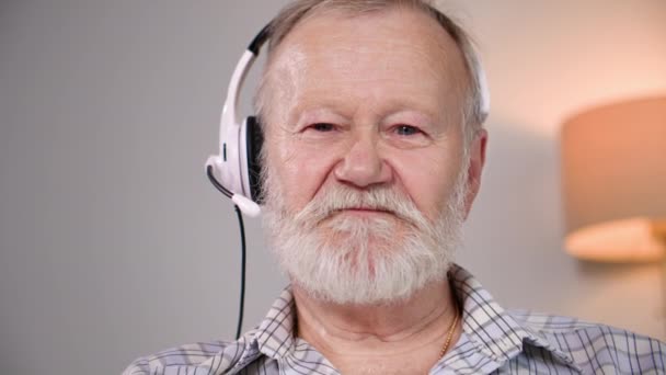 glimlachende man van de pensioengerechtigde leeftijd met een microfoon en hoofdtelefoon communiceert online via videoconferentie op laptop, kijkend naar de camera - Video